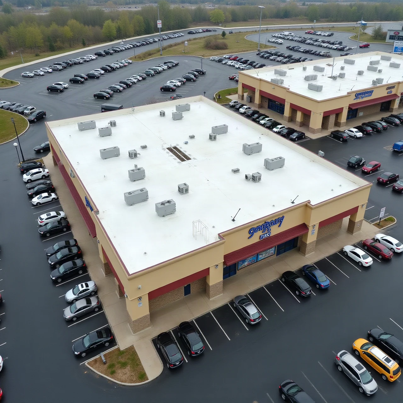 Aerial view of a large single-story retail shopping center with a wide flat low-slope membrane roof, rooftop HVAC units, and a parking lot, overcast daylight, n
